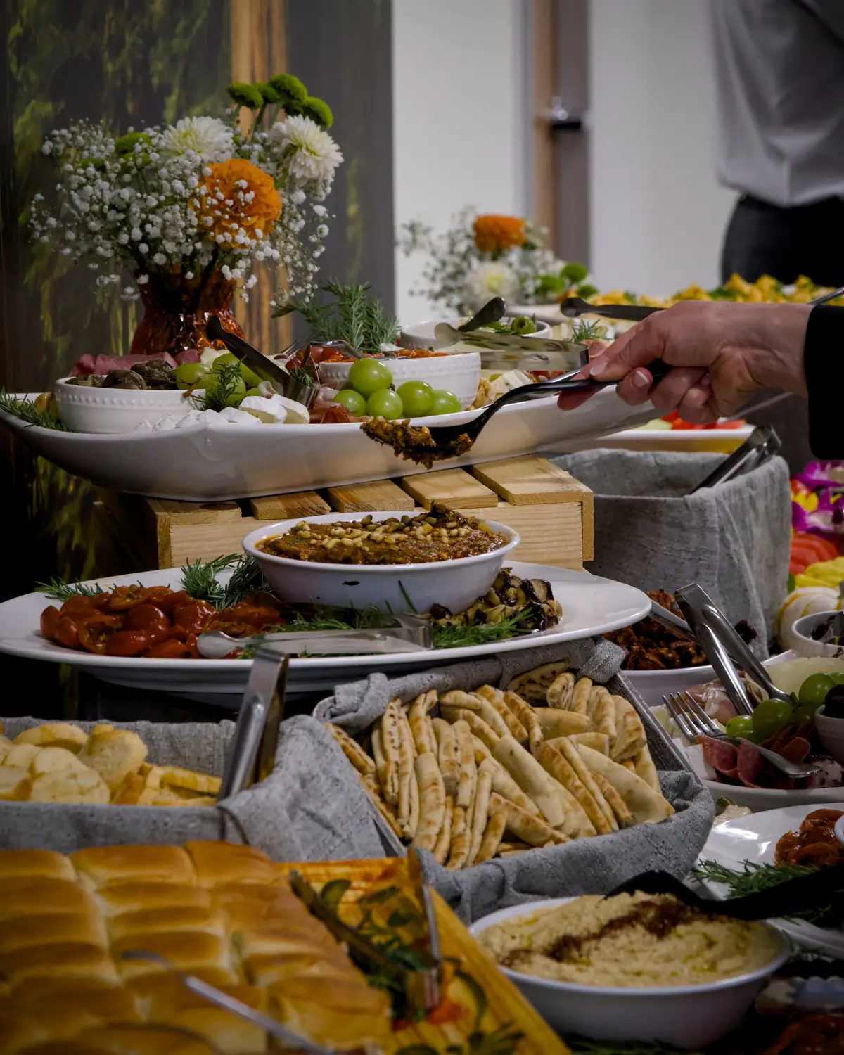A guest serving from a catered spread of cheese, grapes, cured meats, roasted peppers, dips and pita, with flowers behind