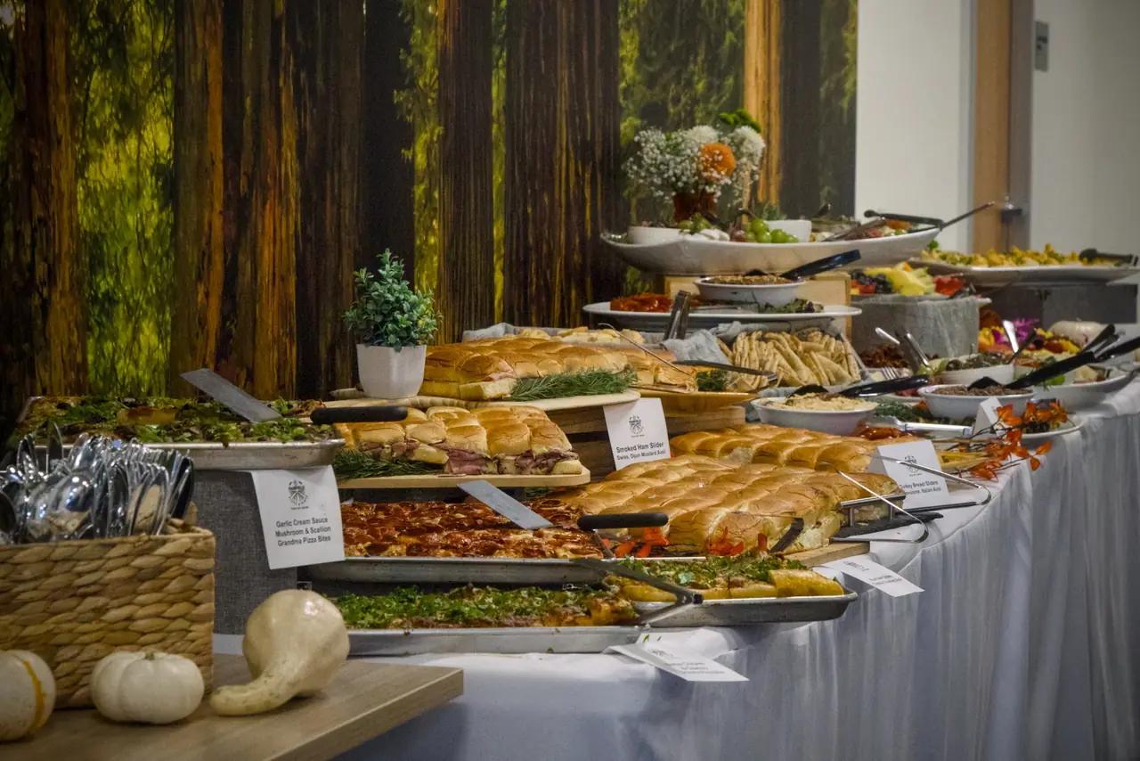 A long catered buffet with labelled trays of grandma pizza and roast beef, smoked ham and turkey sliders, with dips and fruit further down, in front of a forest mural