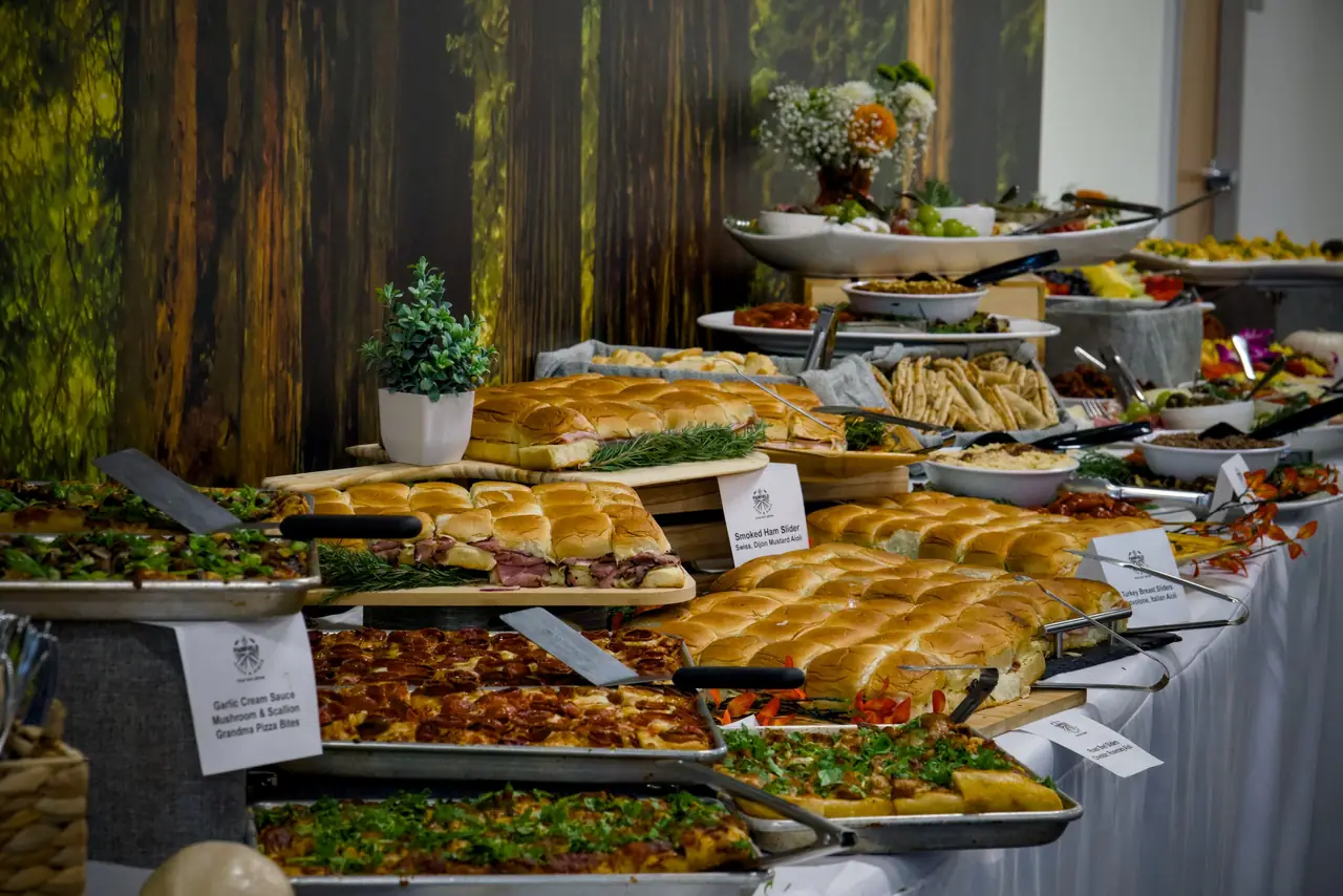 A catered buffet line by Pairfield Pizza & Pints with labelled trays of grandma pizza and sliders in front of a forest mural