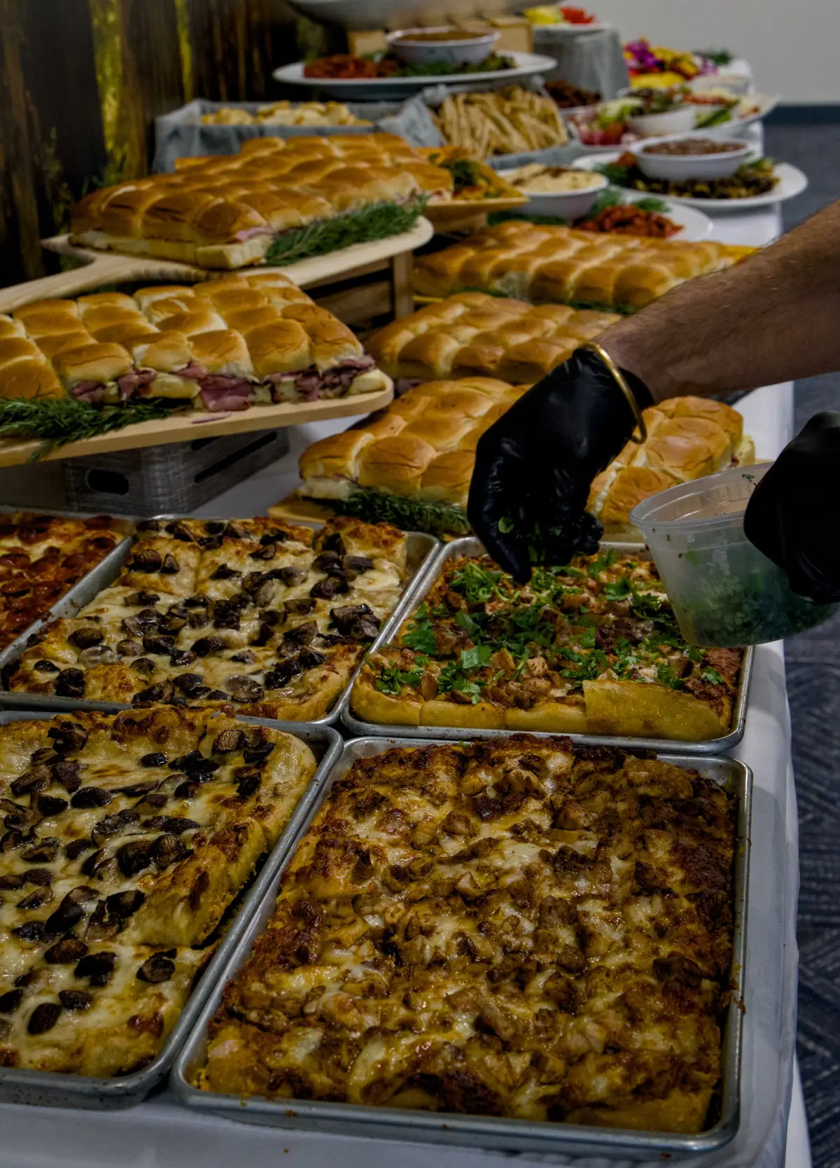 A cook in black gloves scattering cilantro over a tray of butter chicken grandma pizza, next to trays of mushroom grandma pizza and sliders