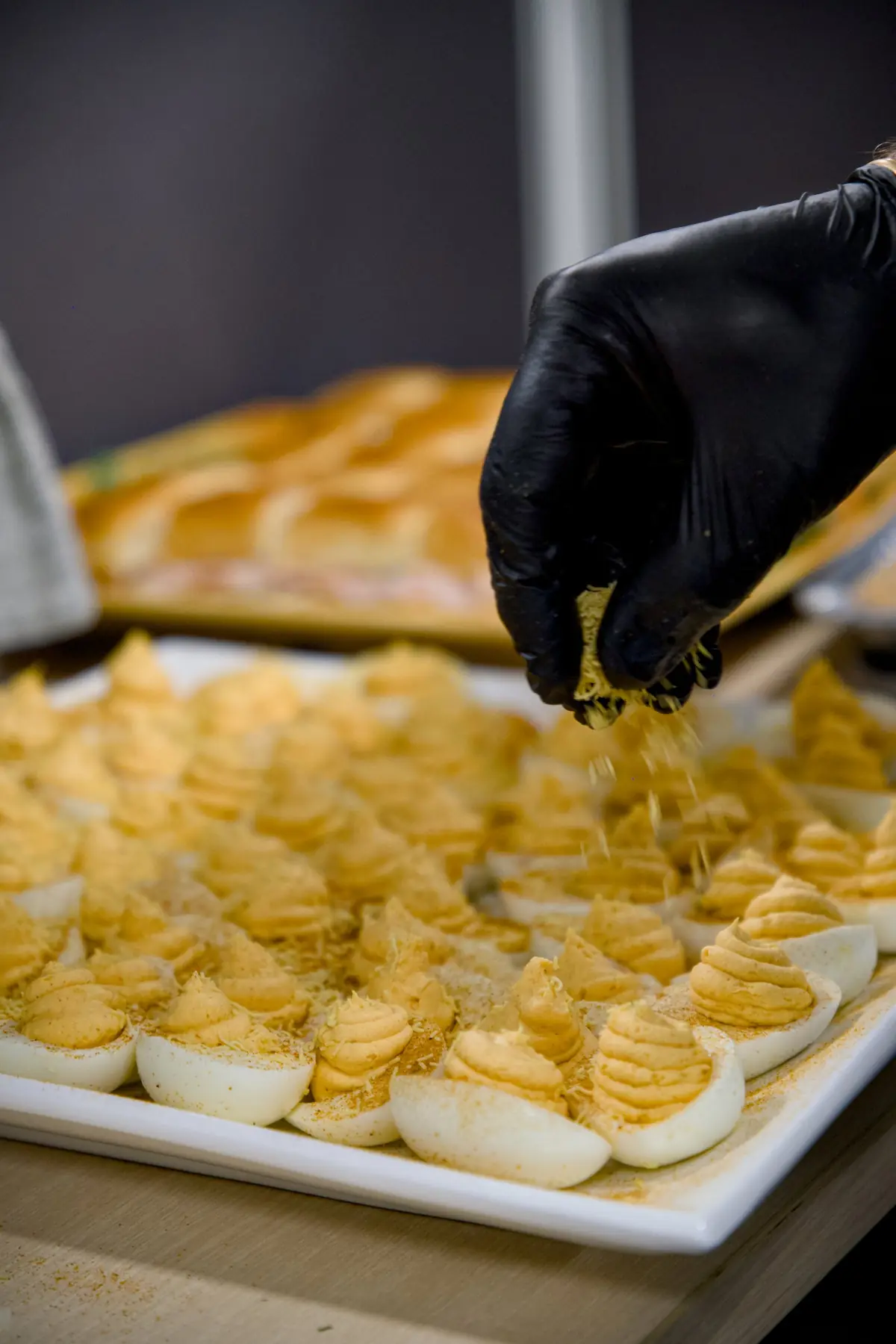 A gloved hand sprinkling a topping over a platter of deviled eggs