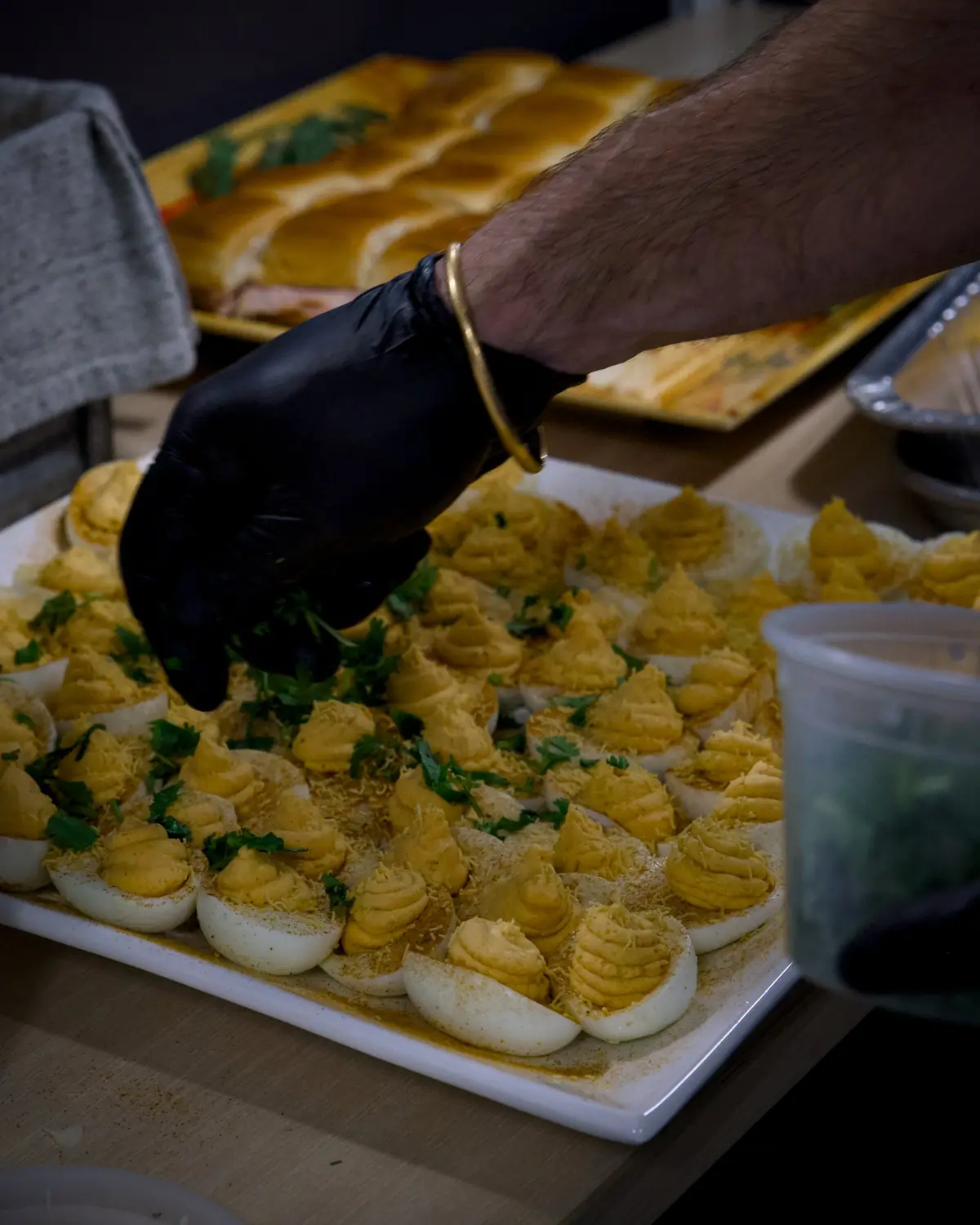 A cook in black gloves garnishing a platter of deviled eggs with cilantro, with a tray of sliders behind