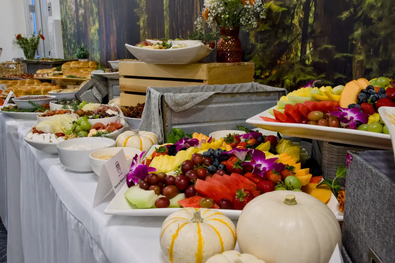 A catered fruit platter of melon, pineapple, strawberries, blueberries, grapes and orchids beside white pumpkins, with cheese and cured meats further along the table