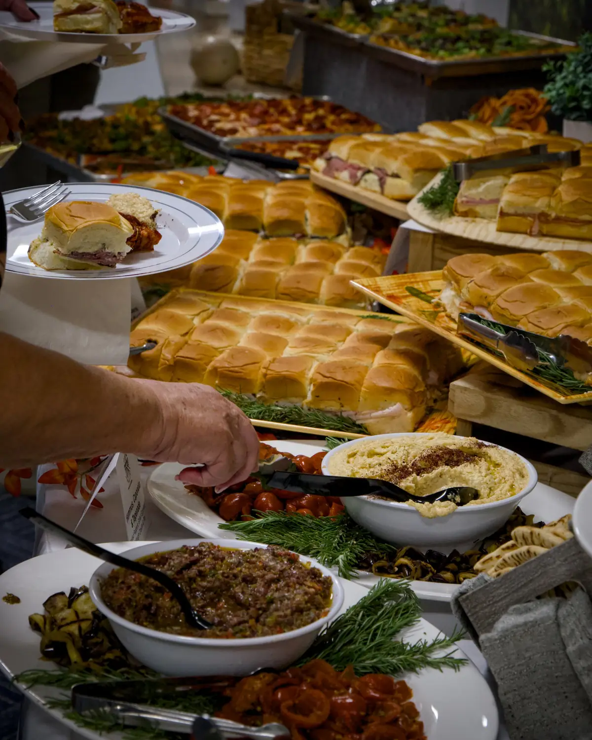 A guest serving roasted peppers beside bowls of hummus and tapenade, in front of trays of sliders
