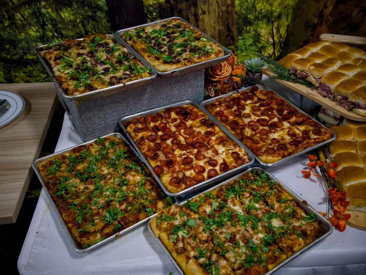 Six trays of mushroom, pepperoni and butter chicken grandma pizza beside roast beef sliders at a catered event