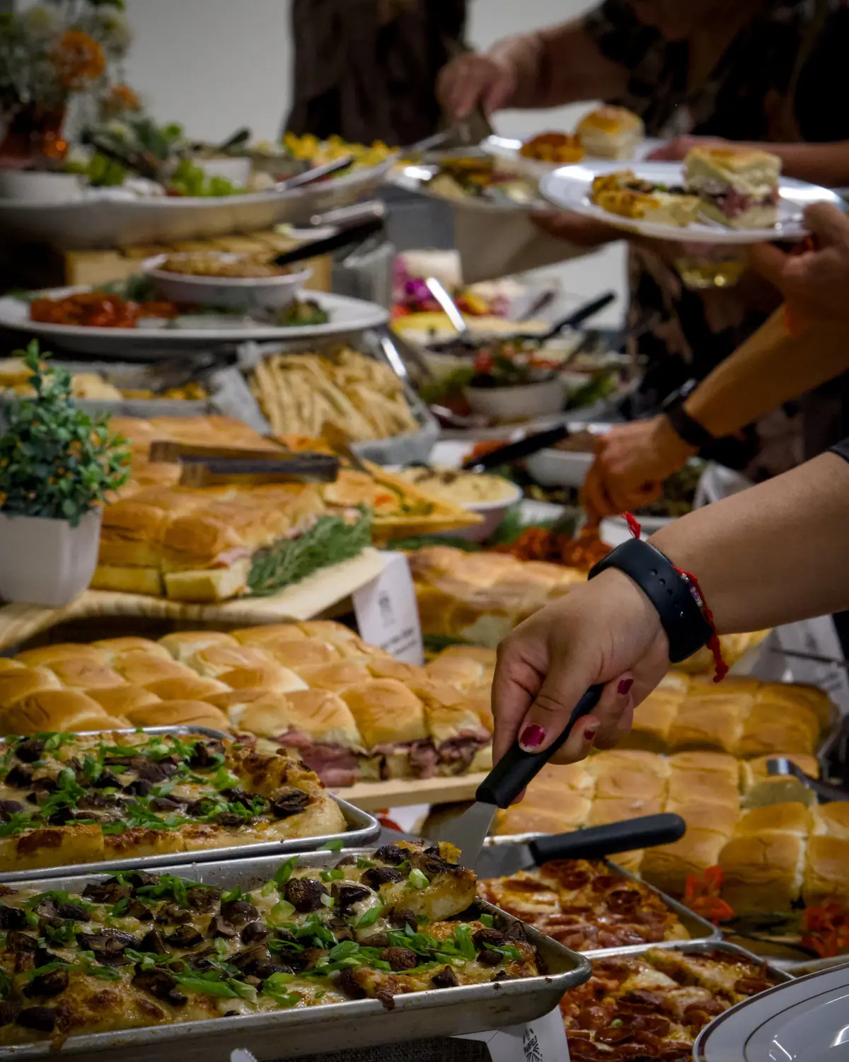 Guests serving themselves at a catered buffet, one lifting a square of mushroom grandma pizza in front of trays of sliders