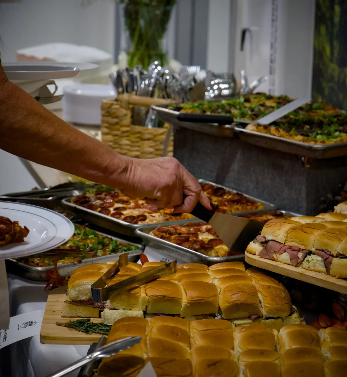A guest lifting a slice of pepperoni grandma pizza at a Pairfield Pizza & Pints catered buffet, beside boards of roast beef sliders
