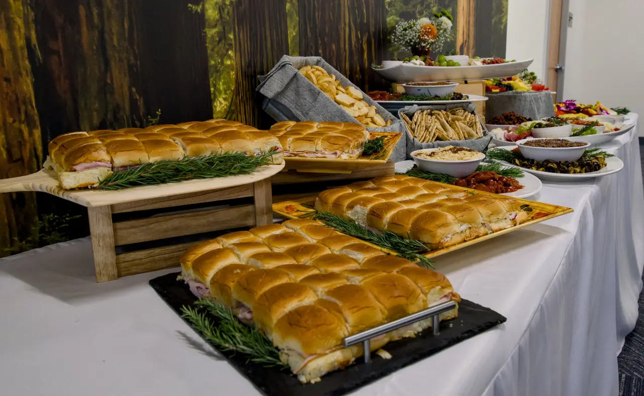 Trays of slider sandwiches garnished with rosemary on a catered buffet, with pita, dips and platters further along