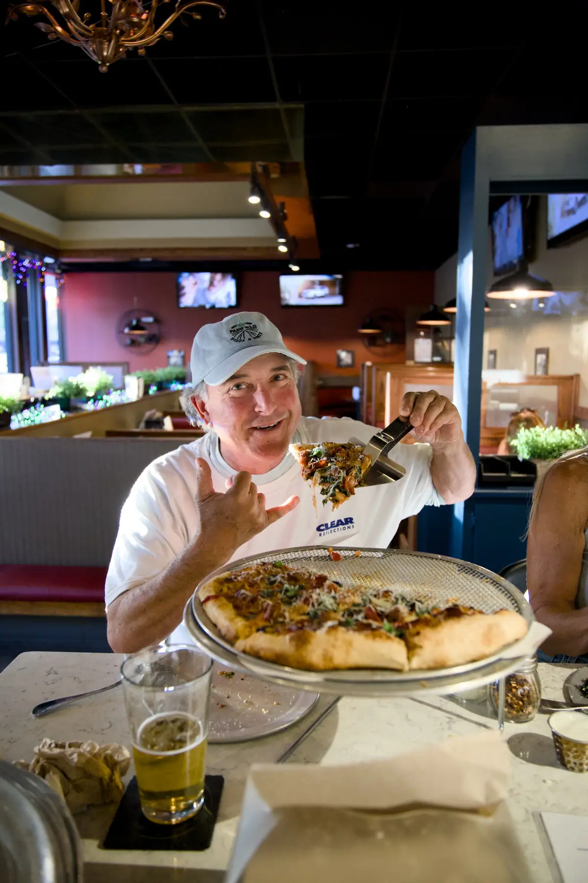 A smiling man in a Pairfield Pizza & Pints cap holding up a slice and giving a shaka, with a pint of beer on the table