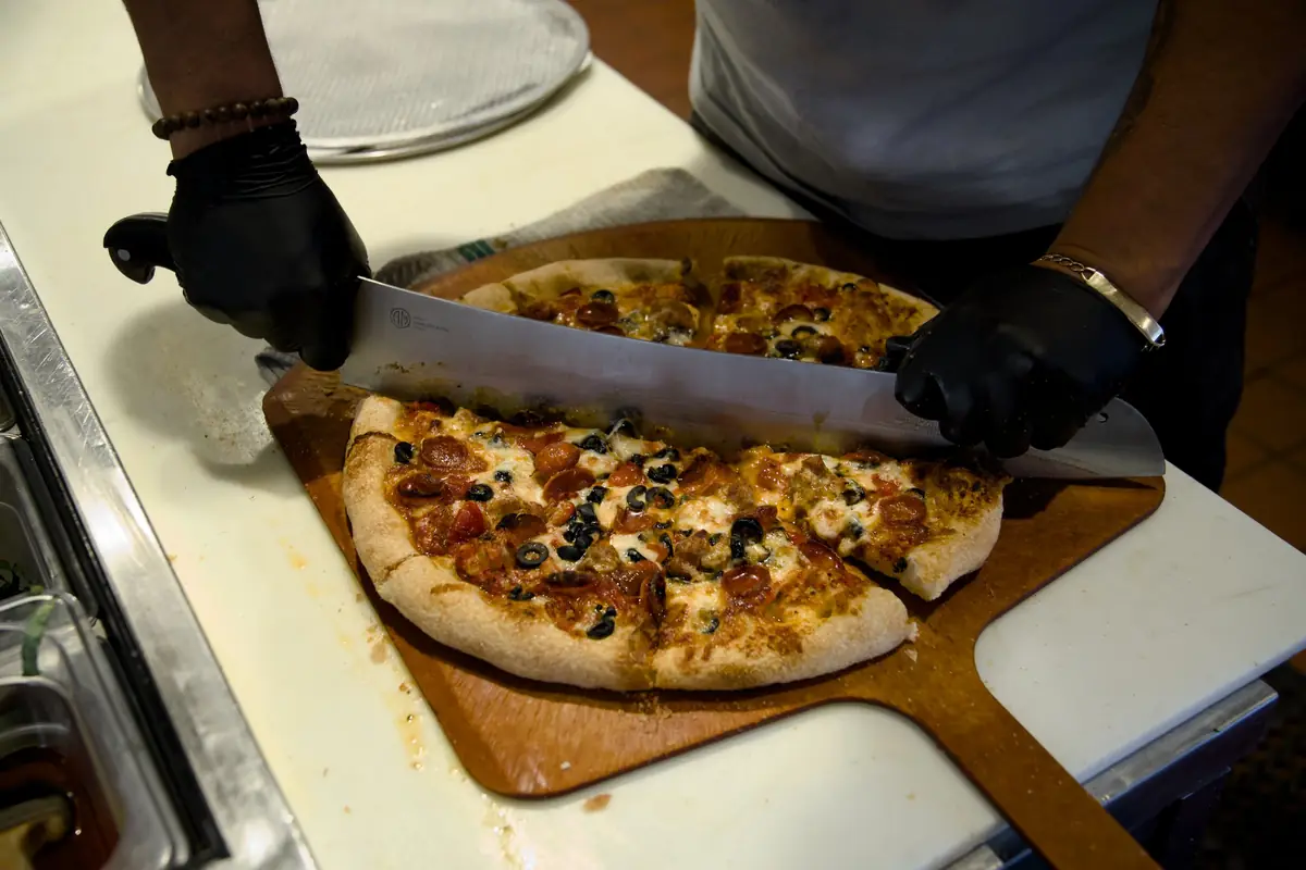 A cook in black gloves slicing a pepperoni, sausage and black olive pizza with a long knife on a wooden peel at Pairfield Pizza & Pints