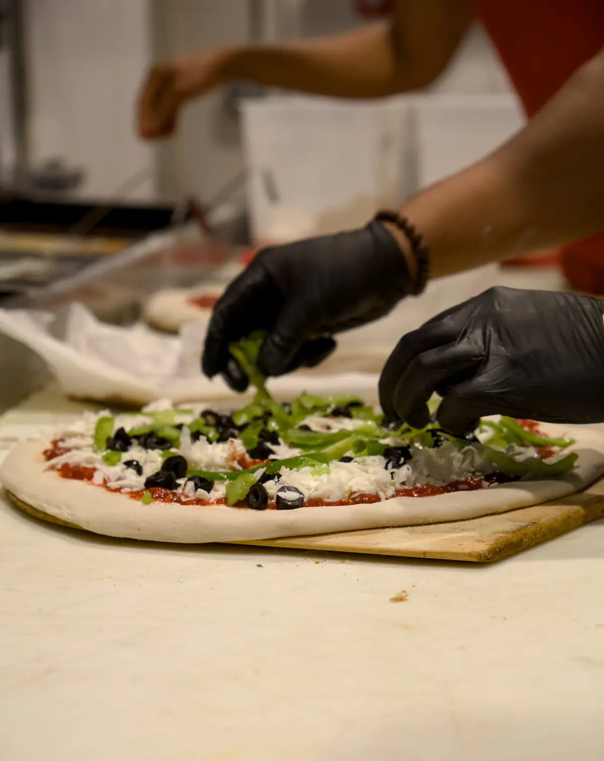 Gloved hands scattering green peppers over a pizza topped with sauce, mozzarella and black olives, ready for the oven at Pairfield Pizza & Pints
